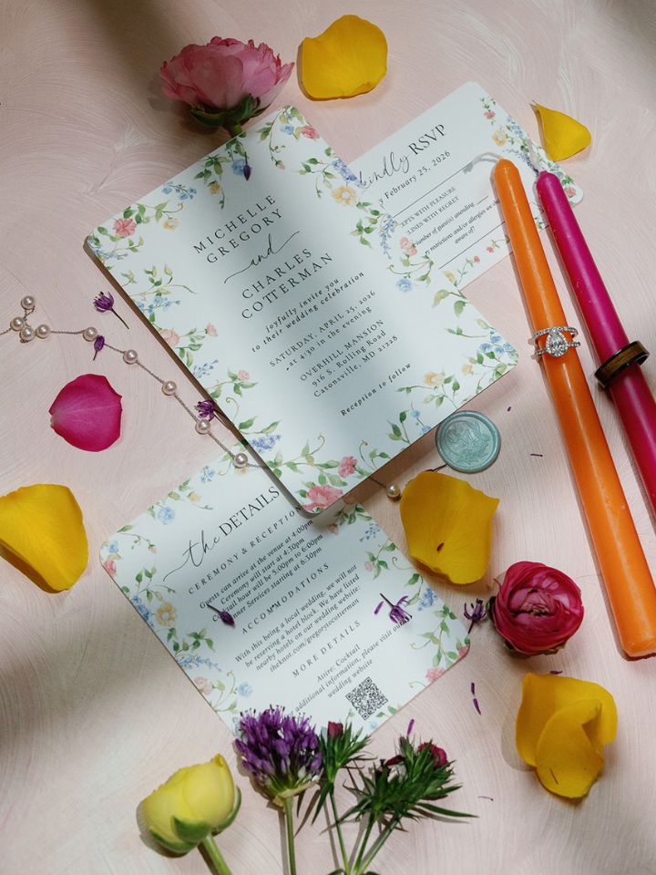 Flat lay photo with wedding invitation, taper candles, rings, and flowers.