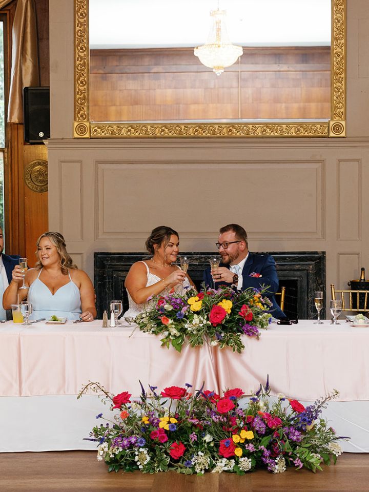 Bride and Groom sit at head table at wedding reception, with large flower arrangements.