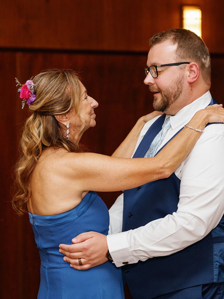 Mother of groom dances with son, wears flower comb in hair.