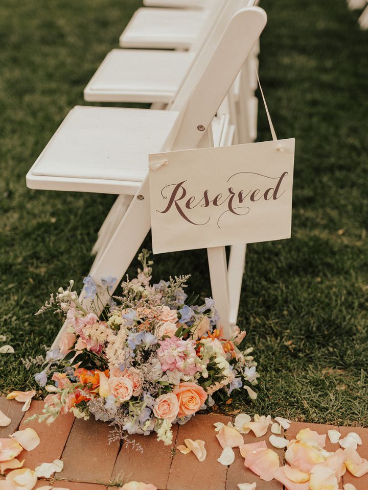 White folding chair at wedding ceremony with reserved sign and flower arrangement at base.