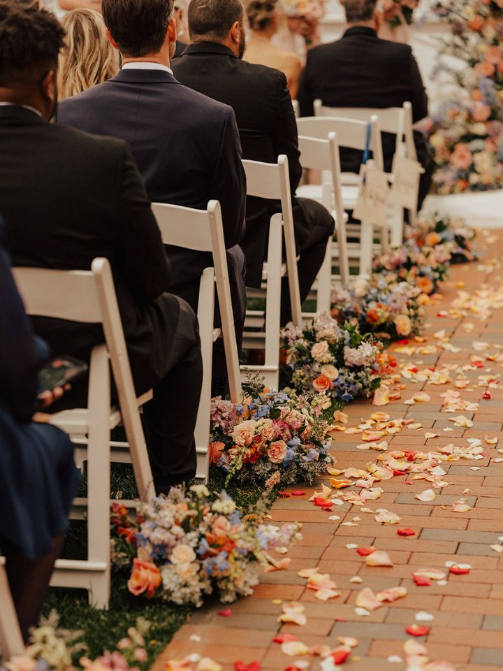 Guests sit in chairs at wedding ceremony, with petals and arrangements lining the sides.
