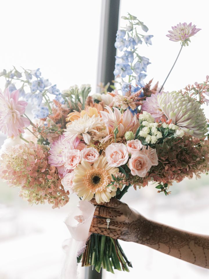 Pastel pink, peach, blue, and yellow flowers in a bridal bouquet, bride's hand holding the bouquet has henna tattoos.