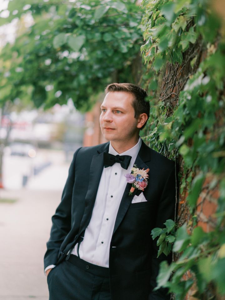 Groom in black tuxedo leans on brick wall with green vines.