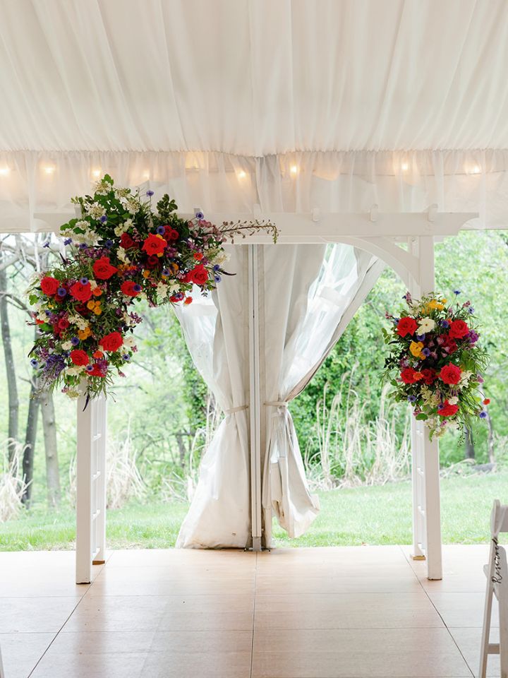 White wedding ceremony pergola underneath white tent with bright spring wedding flower arrangements.