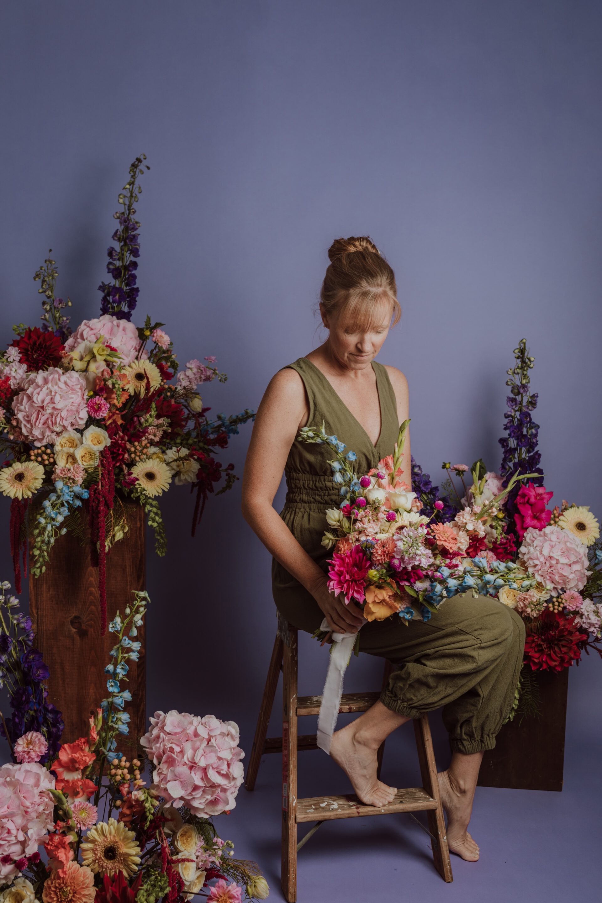 Liza looking down at an arrangement, surrounded by floral arrangements in a studio with a purple backdrop